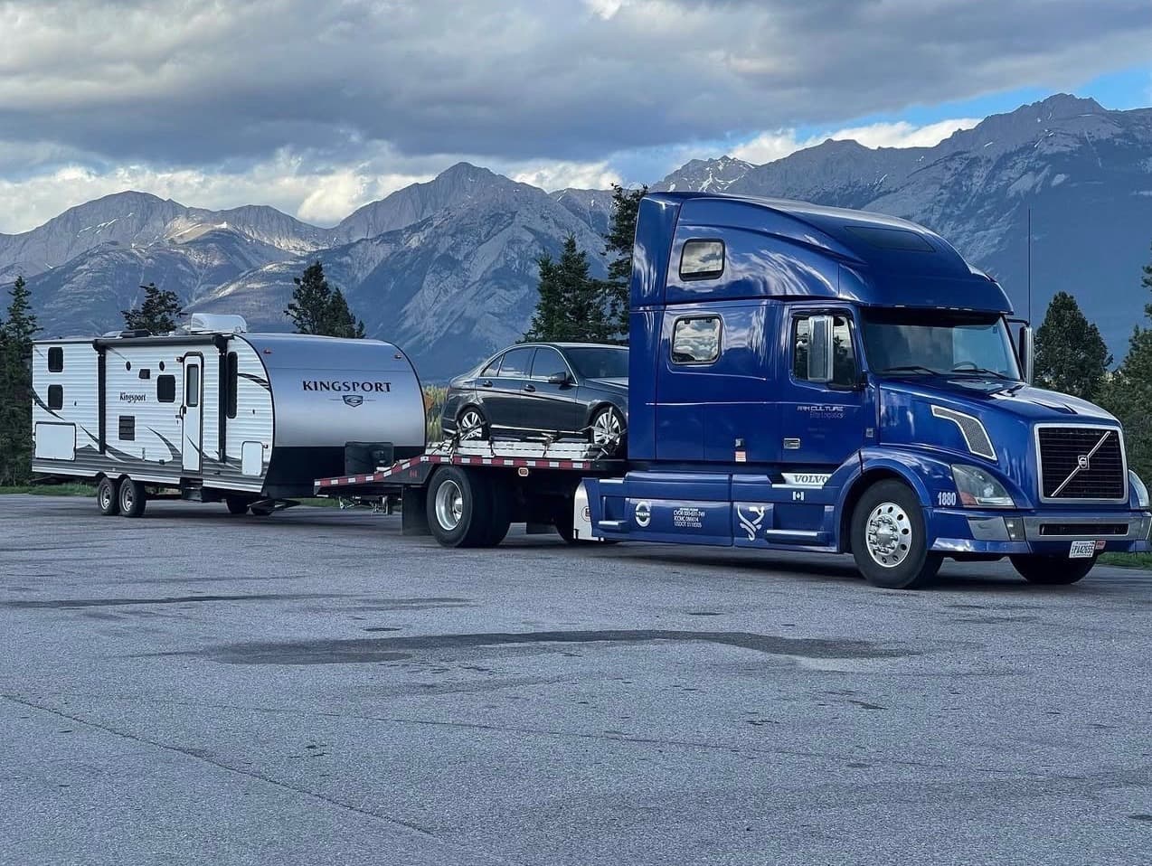 Blue Volvo semi truck hauling a car and a Kingsport travel trailer, parked in front of snow-capped mountains