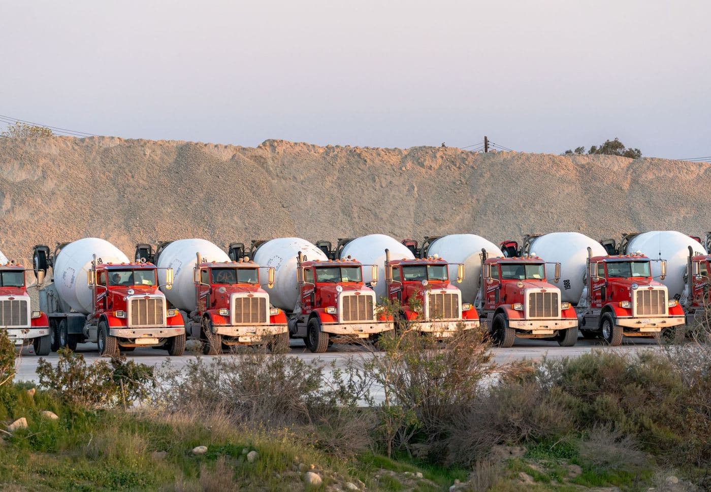 Row of red concrete mixer trucks parked in front of a gravel stockpile