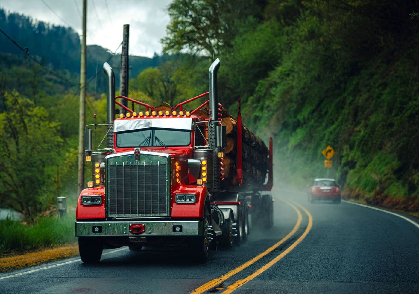 Red long-hood semi truck hauling logs on a wet, winding highway
