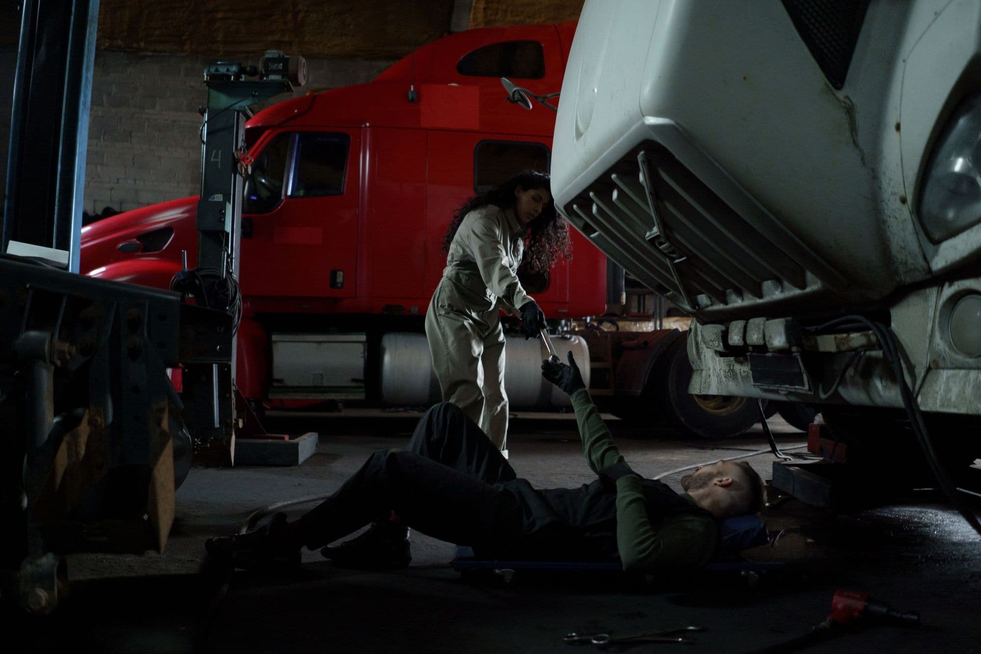 Mechanic lying under a semi truck while a coworker hands him a tool